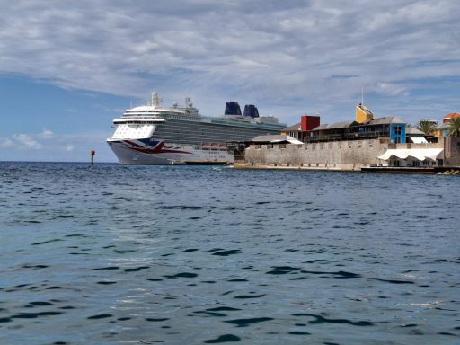 A cruise ship docked at a port beside calm blue waters and a cloudy sky.