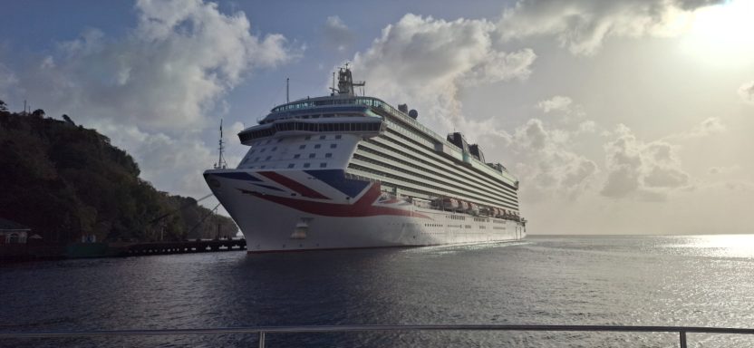 Large cruise ship sails near a coastal landscape under a sunny sky.