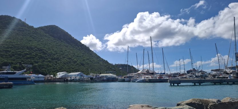 Harbour scene with boats, mountains, and bright blue sky with scattered clouds.