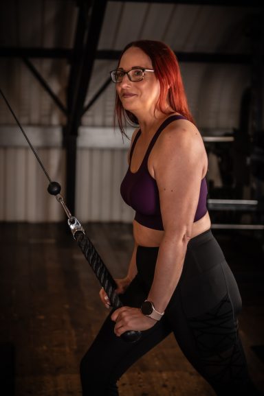 Jodie with red hair in a gym, using a cable machine with focused expression.