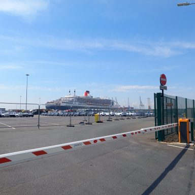 Cruise ship docked near a fenced parking area under a clear blue sky.