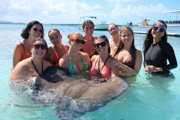 Jodie and her friends in swimwear holding a stingray in shallow water, smiling and enjoying the sun.