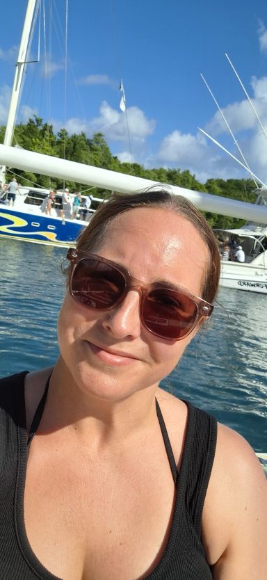 Jodie with sunglasses smiles by the water, with boats in the background and blue sky.