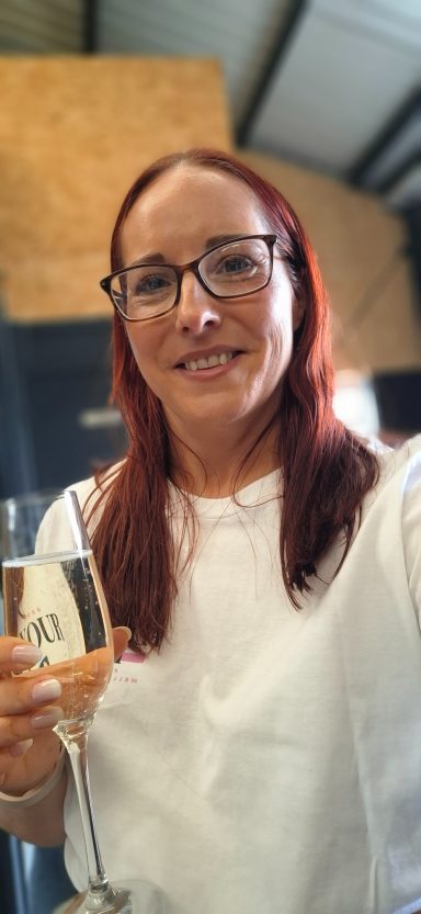 Jodie smiling with red hair, wearing glasses and holding a glass of prosecco.