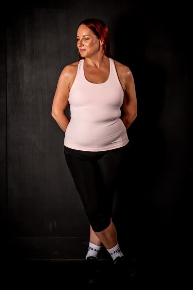 Jodie in a pink tank top and black leggings stands confidently with hands behind her back.