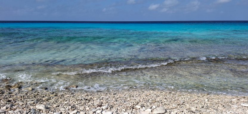 Clear turquoise waves lapping gently on a pebbly beach under a bright sky.
