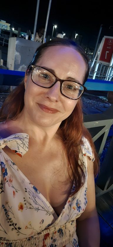 A woman with long hair wearing glasses, smiling against a night city backdrop.