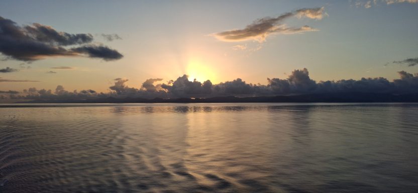 Sunrise over calm water, with clouds reflecting light in soft hues.