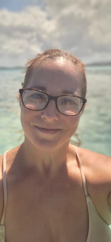 Jodie with glasses smiling in shallow clear water under a blue sky.