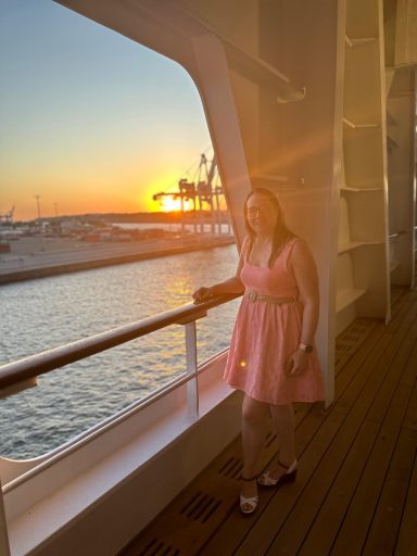 Jodie in a pink dress stands by a railing, with a sunset over a marina in the background.