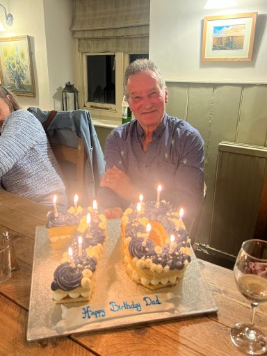 Jodie's Dad smiling sits at a table with his 70th birthday cake adorned with lit candles.
