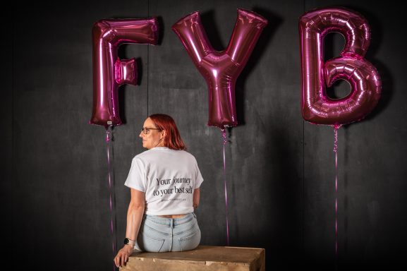 Jodie with red hair sitting on a box, facing away, looking over her left shoulder with pink "FYB" balloons in the background.