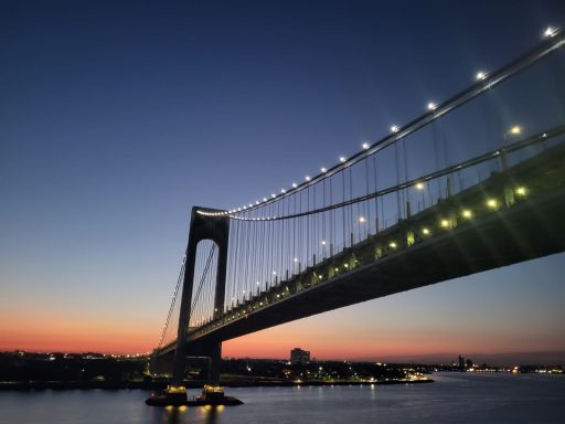 Suspension bridge at sunset, with illuminated cables reflecting on the water below.
